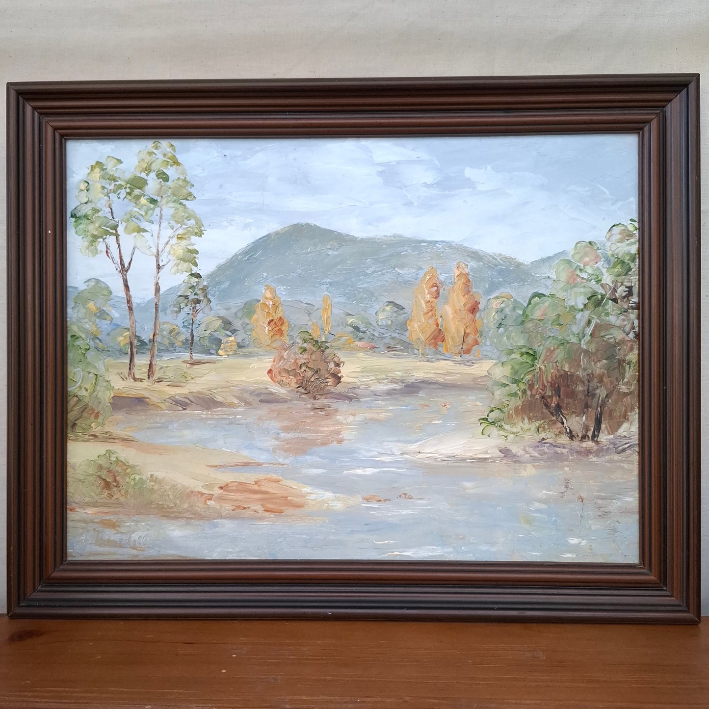 Framed painting of a landscape with trees and mountains on a wall.