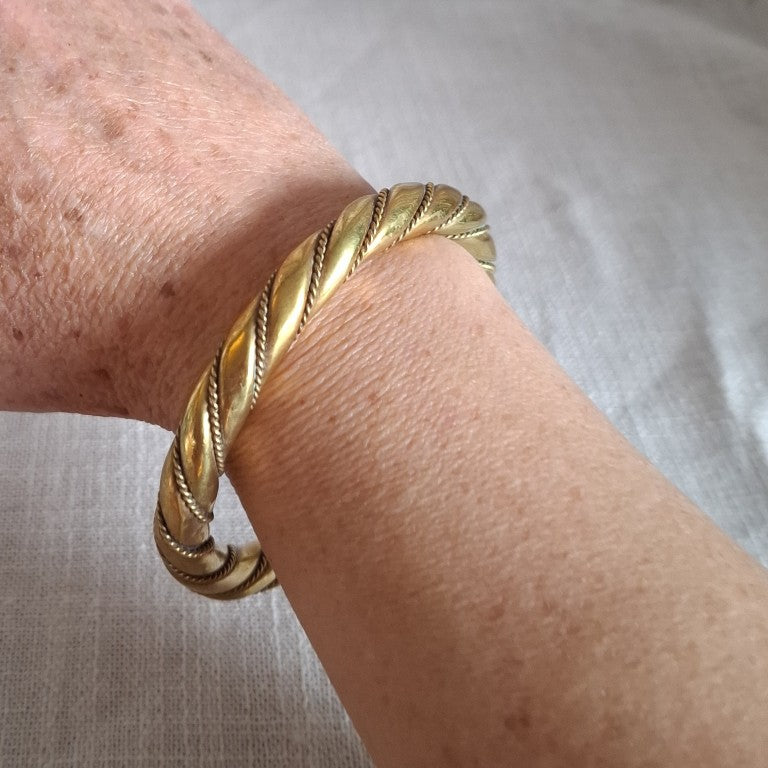 A close-up of a gold-toned twisted rope cuff bracelet worn on a person's wrist against a light fabric background.